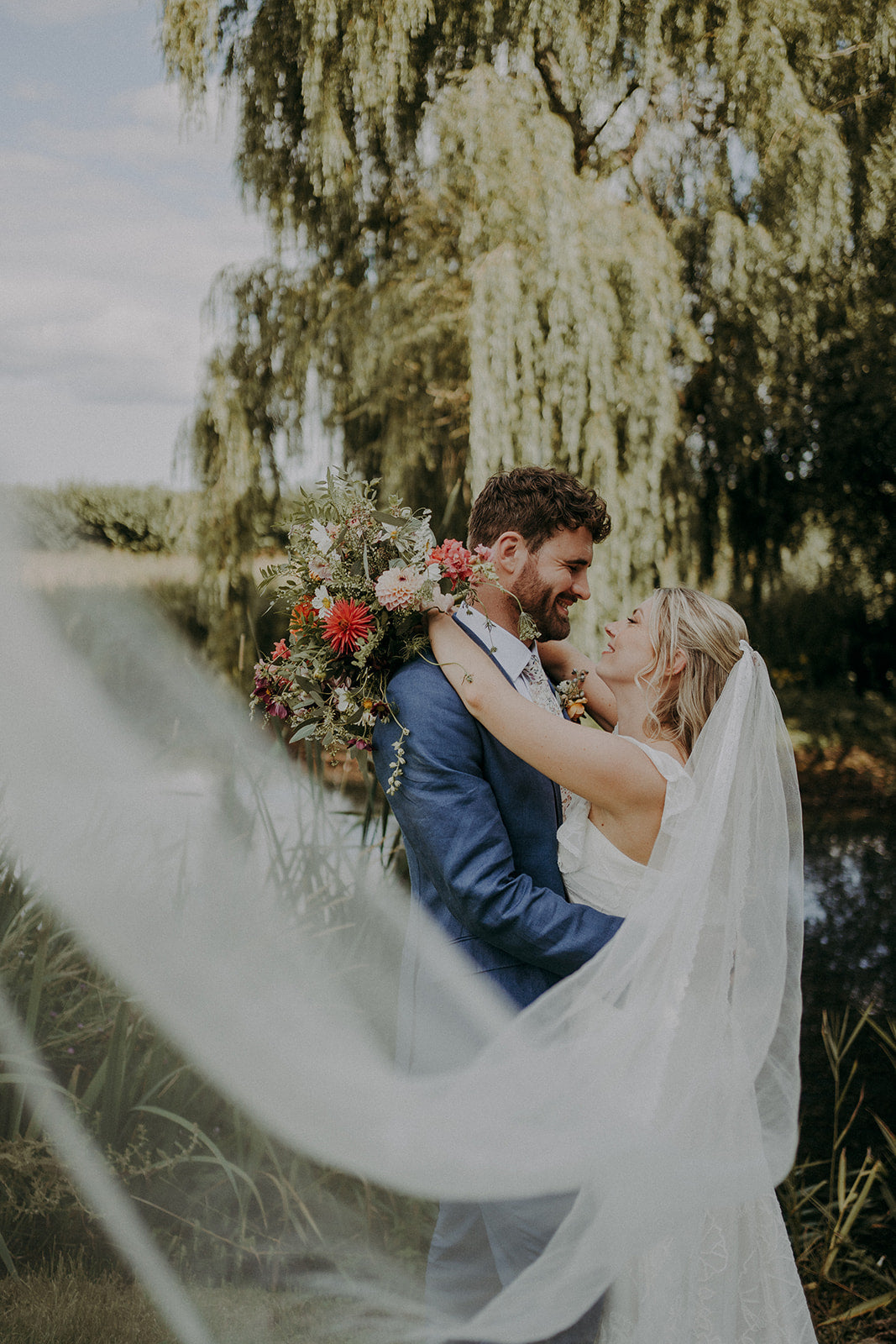 Bohemian bride and groom embracing each other in front of trees and a lake.