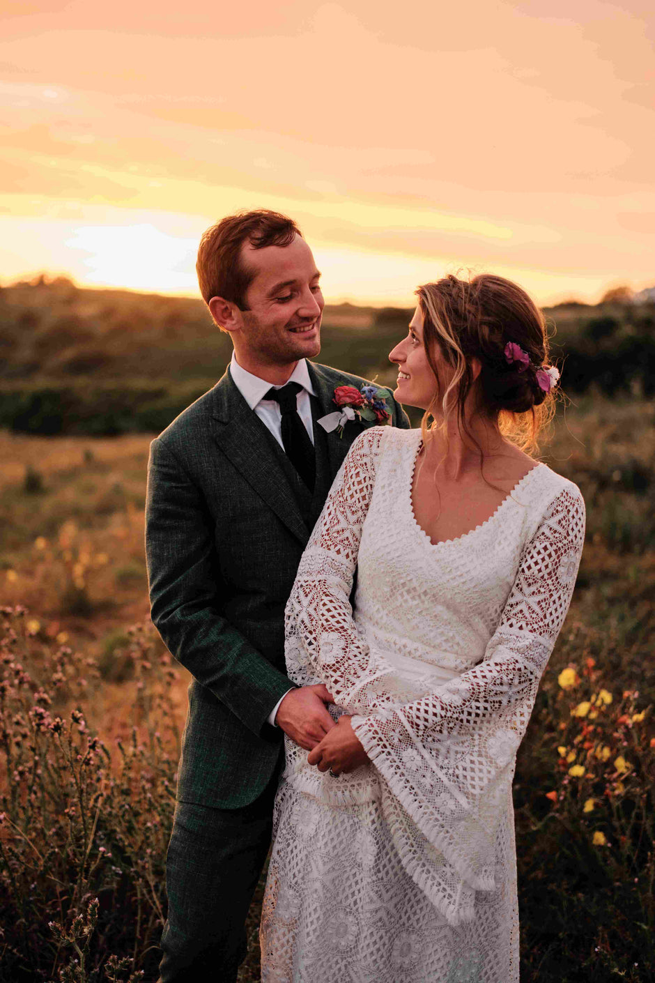 bride wearing a bohemian lace wedding dress with bell sleeves standing with the groom in sunset